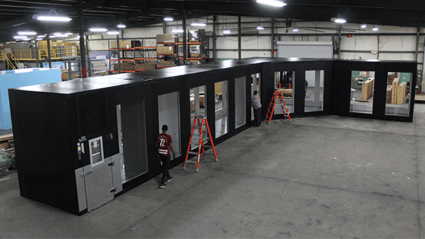 View of Thermo-Kool factory floor with L shaped walk-in unit being installed. Two orange ladders and a worker with back to the camera are in front of the unit.