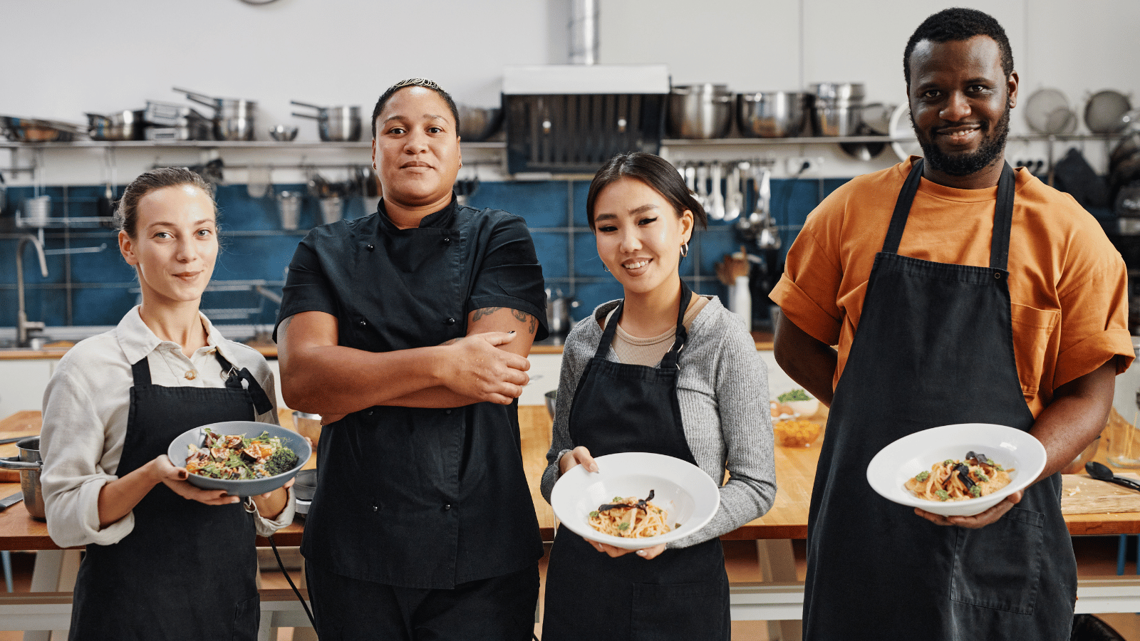 Four workers in a commercial kitchen standing. Three holding white bowls. One in black chef's jacket has arms crossed.