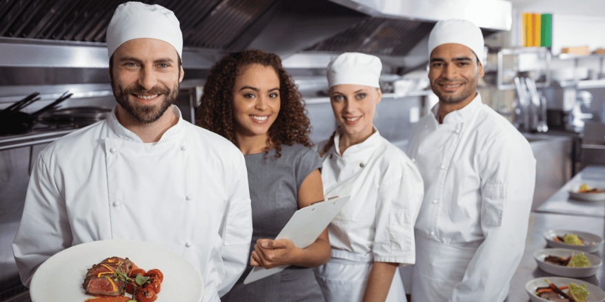 Four workers, three in chef whites and hats and one in grey dress holding pad, in commercial kitchen.
